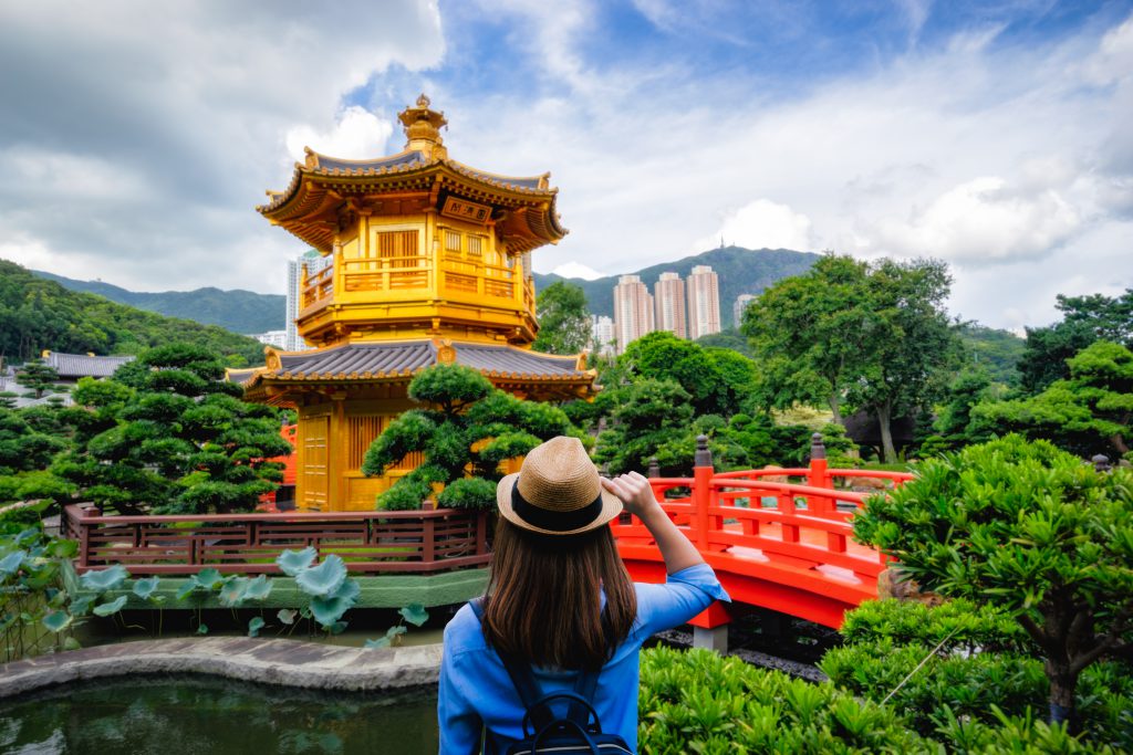 ผู้หญิงสวมหมวกยืนหันหน้าหาเจดีย์สถาปัตกรรมจีนสีเหลืองทอง ด้านข้างมีสะพานไม้สีแดง